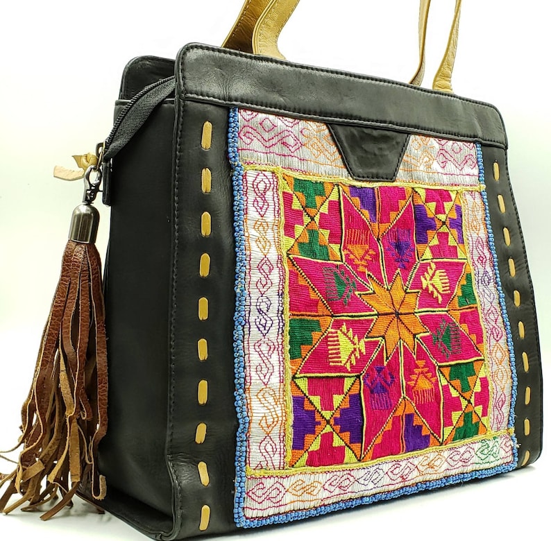 May include: Black leather tote bag with a colourful embroidered panel featuring a starburst design in pink, orange, yellow, and green. The bag has gold-coloured handles and a brown leather tassel. The stitching is accented with gold thread.