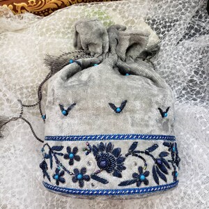 May include: A gray velvet drawstring bag with blue and silver embroidery. The bag has a tassel and is decorated with blue beads.