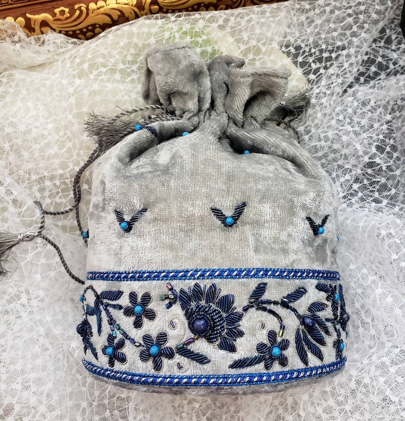 May include: A gray velvet drawstring bag with blue and silver embroidery. The bag has a tassel and is decorated with blue beads.