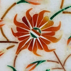 Puede incluir: Primer plano de una incrustaci&oacute;n de m&aacute;rmol blanco con un dise&ntilde;o floral rojo, naranja y verde. La flor est&aacute; en el centro de la imagen y tiene una forma circular con p&eacute;talos que irradian hacia afuera. La flor est&aacute; rodeada de hojas y tallos.