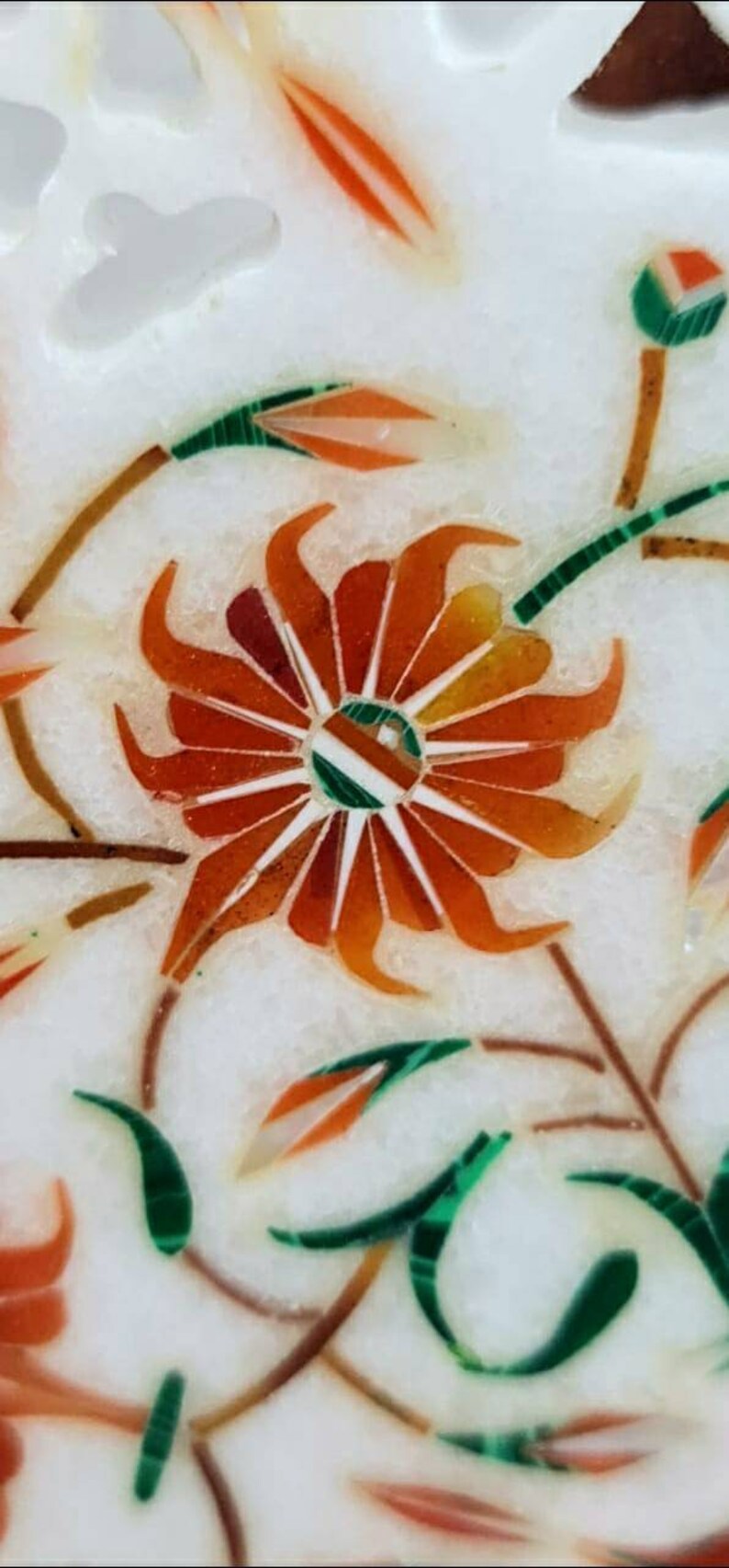 Puede incluir: Primer plano de una incrustaci&oacute;n de m&aacute;rmol blanco con un dise&ntilde;o floral rojo, naranja y verde. La flor est&aacute; en el centro de la imagen y tiene una forma circular con p&eacute;talos que irradian hacia afuera. La flor est&aacute; rodeada de hojas y tallos.