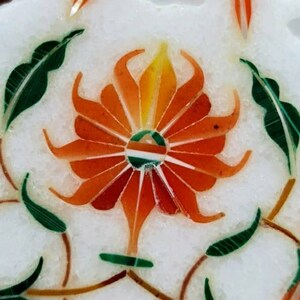 Puede incluir: Primer plano de una incrustaci&oacute;n de m&aacute;rmol blanco con un dise&ntilde;o floral. La flor es naranja y amarilla con hojas verdes. El centro de la flor es un c&iacute;rculo con un dise&ntilde;o verde, blanco y naranja.