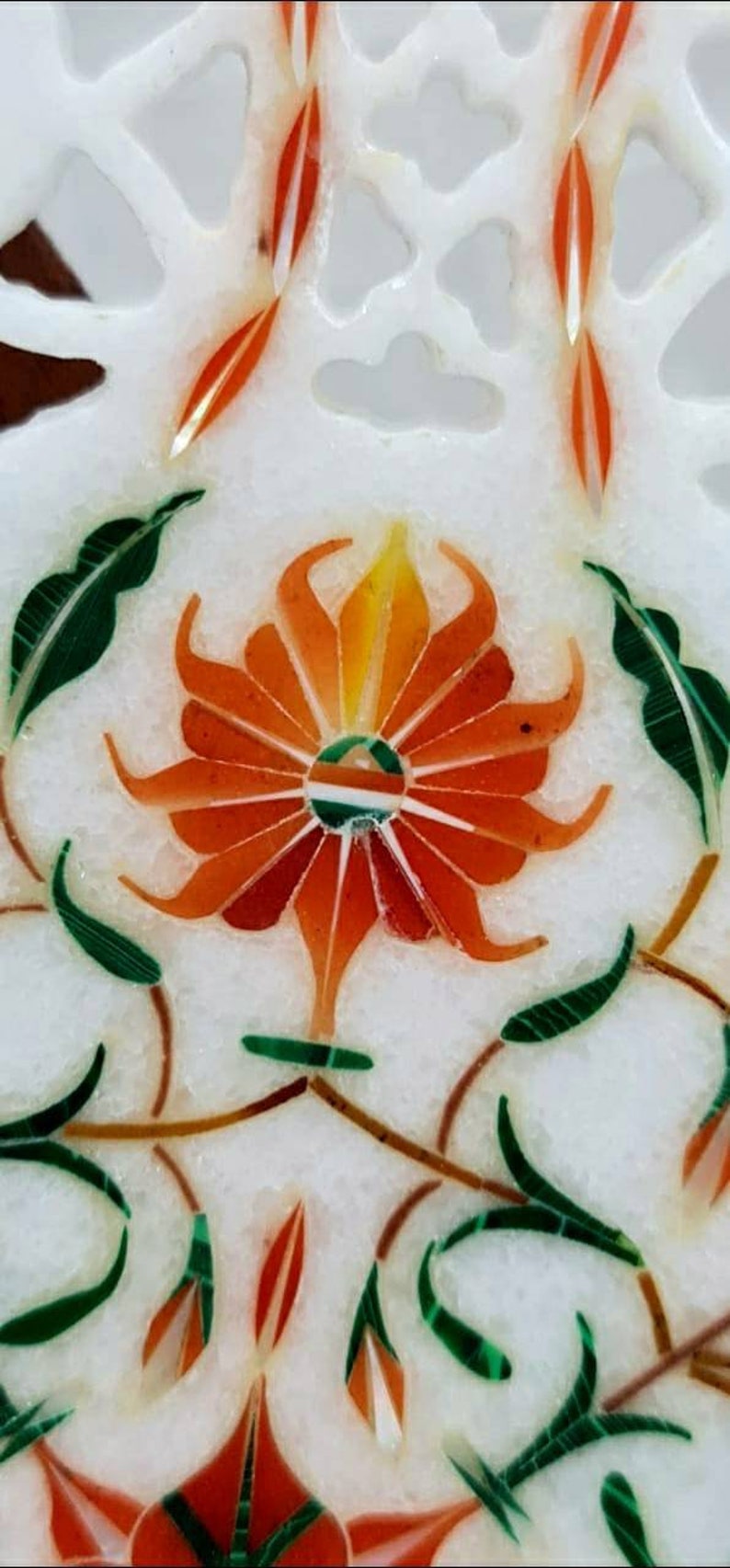 Puede incluir: Primer plano de una incrustaci&oacute;n de m&aacute;rmol blanco con un dise&ntilde;o floral. La flor es naranja y amarilla con hojas verdes. El centro de la flor es un c&iacute;rculo con un dise&ntilde;o verde, blanco y naranja.