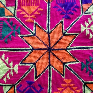 May include: Close-up of a vibrant textile with a geometric pattern. The design features a central orange star surrounded by pink, green, purple, and yellow shapes. The stitching is visible, creating a textured surface.