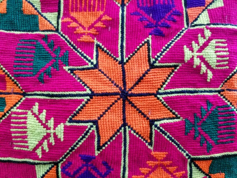 May include: Close-up of a vibrant textile with a geometric pattern. The design features a central orange star surrounded by pink, green, purple, and yellow shapes. The stitching is visible, creating a textured surface.