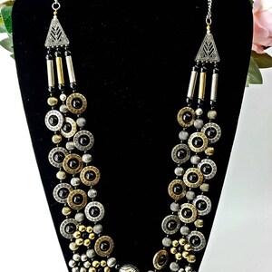 May include: A multi-strand necklace with black, gold, and silver beads. The necklace features a geometric pendant at the top and a variety of round and oval beads.