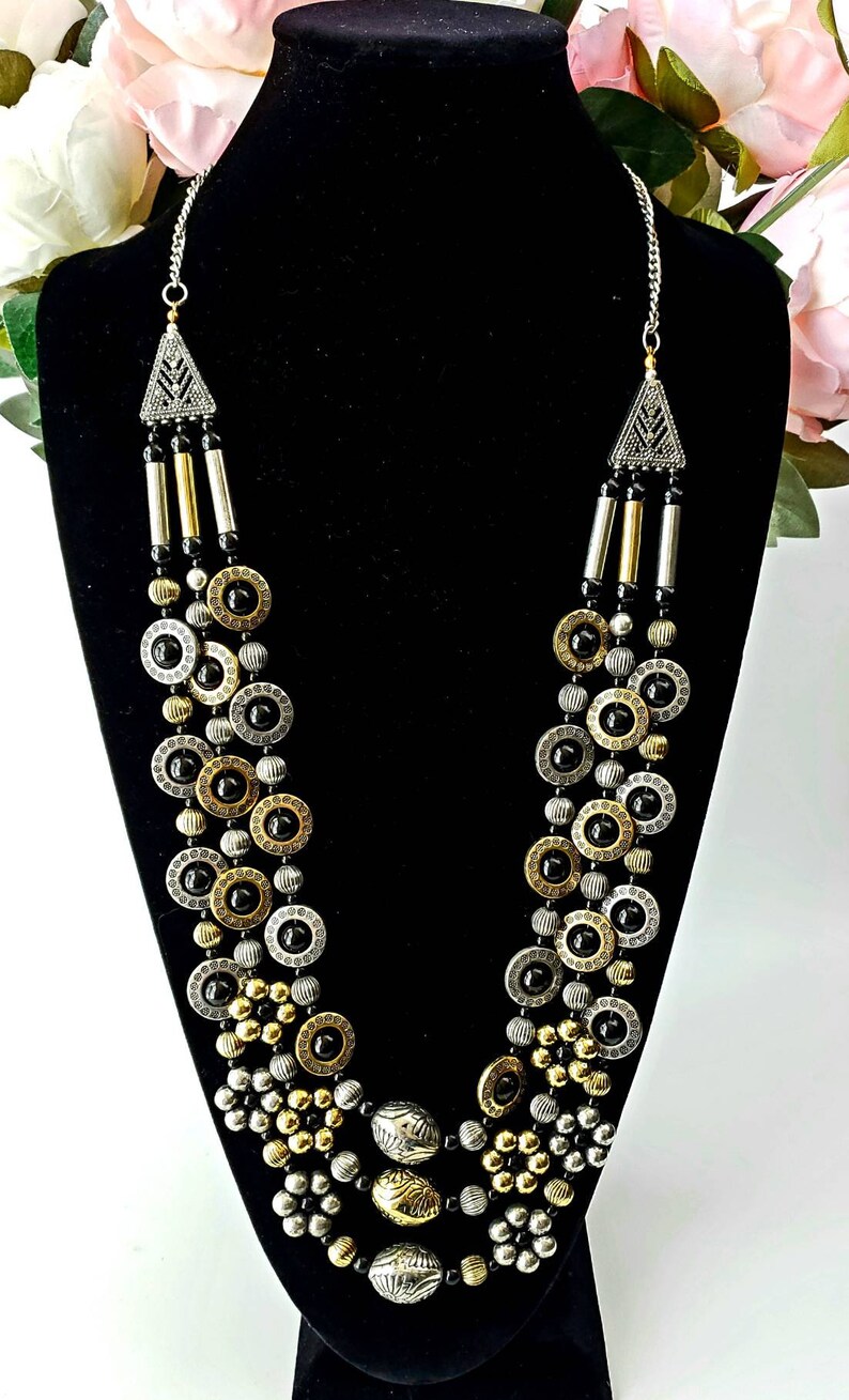 May include: A multi-strand necklace with black, gold, and silver beads. The necklace features a geometric pendant at the top and a variety of round and oval beads.