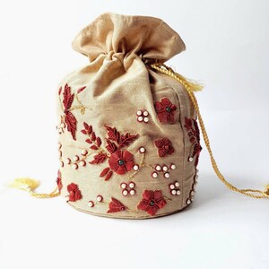 Op de afbeelding: Een beige trekkoordtasje met ingewikkelde rode bloemenborduurwerk en parelaccenten. De tas heeft een gouden trekkoordsluiting en kwastjes, wat een elegant en decoratief ontwerp biedt.