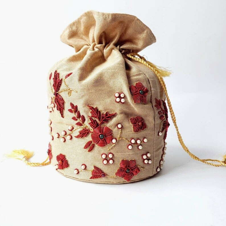 Op de afbeelding: Een beige trekkoordtasje met ingewikkelde rode bloemenborduurwerk en parelaccenten. De tas heeft een gouden trekkoordsluiting en kwastjes, wat een elegant en decoratief ontwerp biedt.