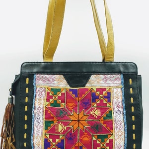 May include: Black leather tote bag with tan leather handles and a colourful embroidered front panel. The panel features a geometric starburst design in shades of pink, orange, purple, and green. A brown tassel hangs from the side.