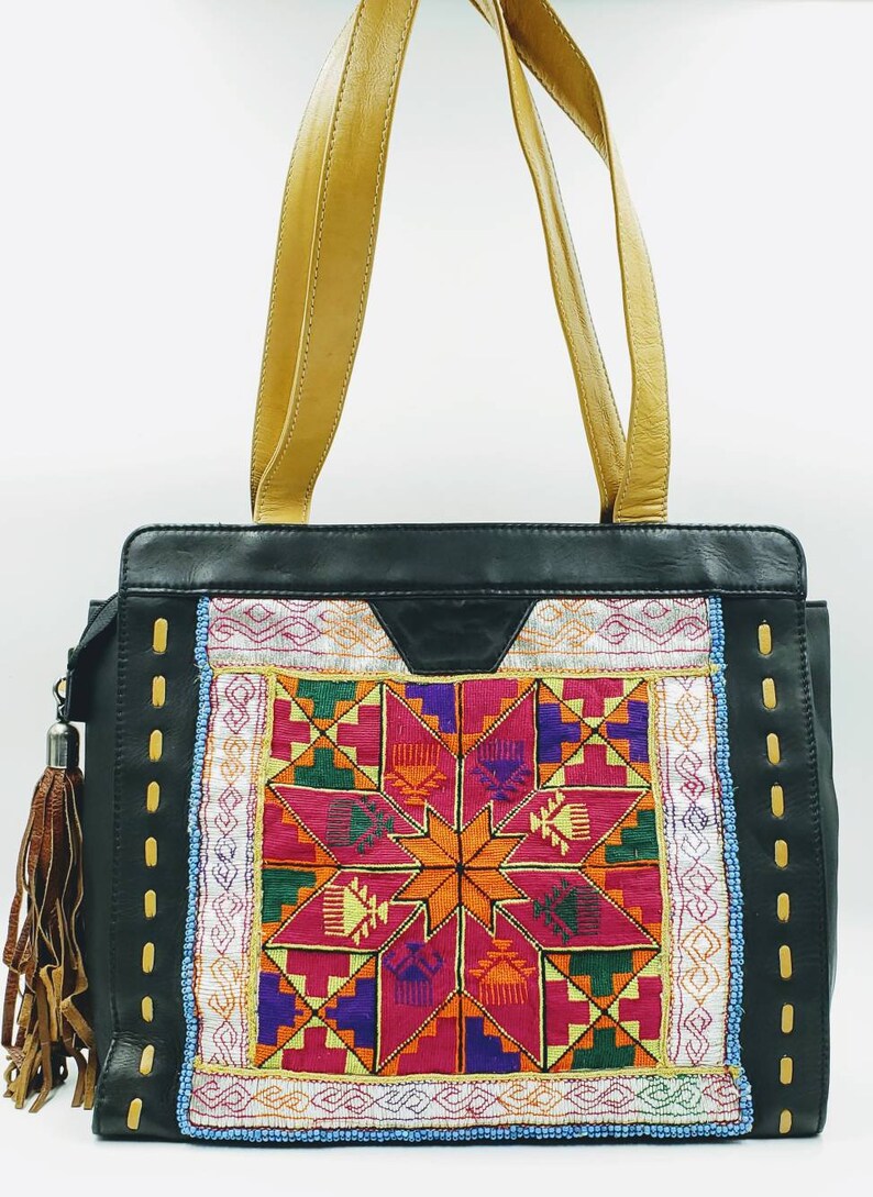 May include: Black leather tote bag with tan leather handles and a colourful embroidered front panel. The panel features a geometric starburst design in shades of pink, orange, purple, and green. A brown tassel hangs from the side.