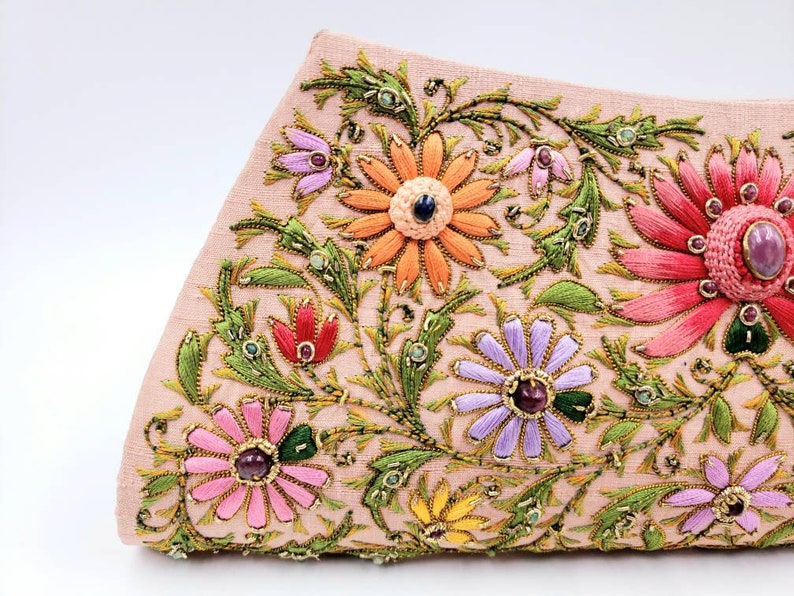 May include: A pink fabric clutch bag with embroidered floral designs in shades of pink, orange, yellow, green, and purple. The flowers have jeweled centers.