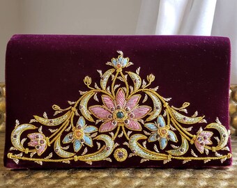 Könnte beinhalten: Rechteckige, weinrote Samt-Clutch mit aufwendigem floralem Stickdesign. Die Stickerei weist goldfarbene Fäden, rosa, blaue und grüne Akzente sowie kleine, farbige Edelsteine auf. Die Clutch ist geschlossen und liegt auf einer goldenen Oberfläche.