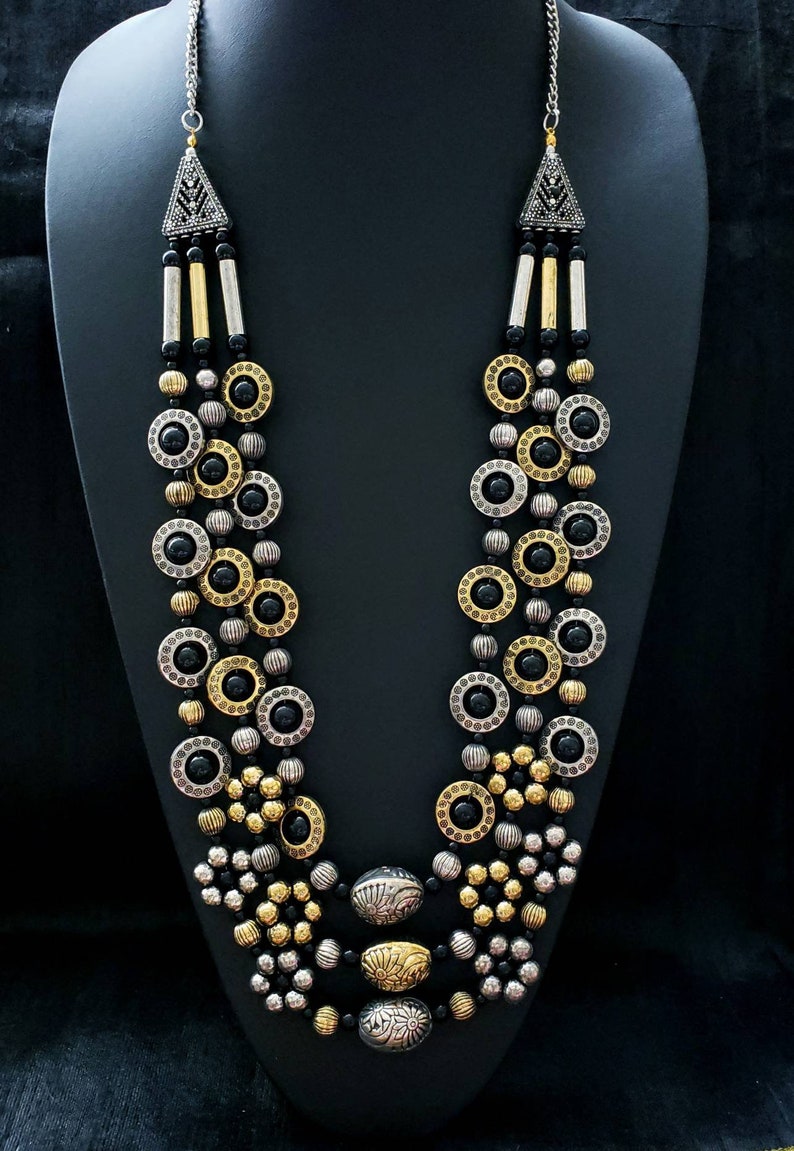 May include: A multi-strand necklace with black, gold, and silver beads. The necklace features round, patterned beads and a large, ornate pendant.