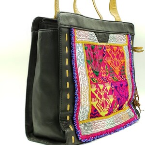 May include: Black tote bag with a colourful embroidered panel featuring pink, yellow, and orange geometric designs. The bag has tan handles and decorative stitching with gold accents. The bag is made of leather.