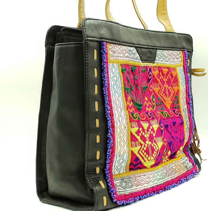 May include: Black tote bag with a colourful embroidered panel featuring pink, yellow, and orange geometric designs. The bag has tan handles and decorative stitching with gold accents. The bag is made of leather.
