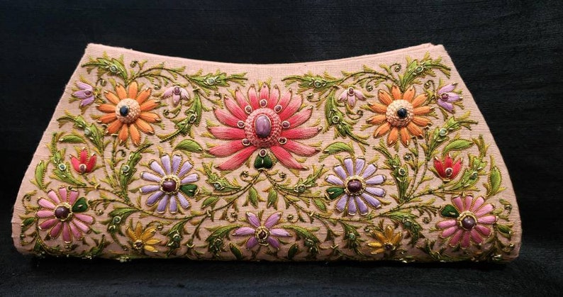 May include: A light pink clutch purse with floral embroidery. The flowers are in shades of pink, orange, purple, and green. The purse is decorated with gold thread and small, colorful beads.
