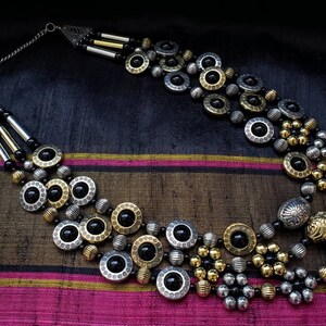 May include: A multi-strand necklace with black, gold, and silver beads. The necklace features large, round, silver and gold beads with black accents. The necklace is on a dark background.