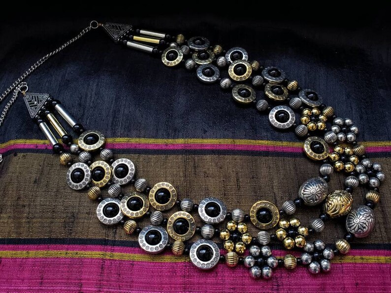 May include: A multi-strand necklace with black, gold, and silver beads. The necklace features large, round, silver and gold beads with black accents. The necklace is on a dark background.