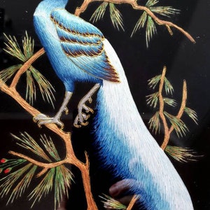 Puede incluir: Un p&aacute;jaro bordado azul y blanco con cabeza roja posado en una rama dorada con agujas de pino verdes. El p&aacute;jaro est&aacute; rodeado de un fondo negro.