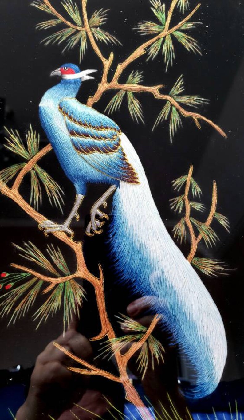 Puede incluir: Un p&aacute;jaro bordado azul y blanco con cabeza roja posado en una rama dorada con agujas de pino verdes. El p&aacute;jaro est&aacute; rodeado de un fondo negro.