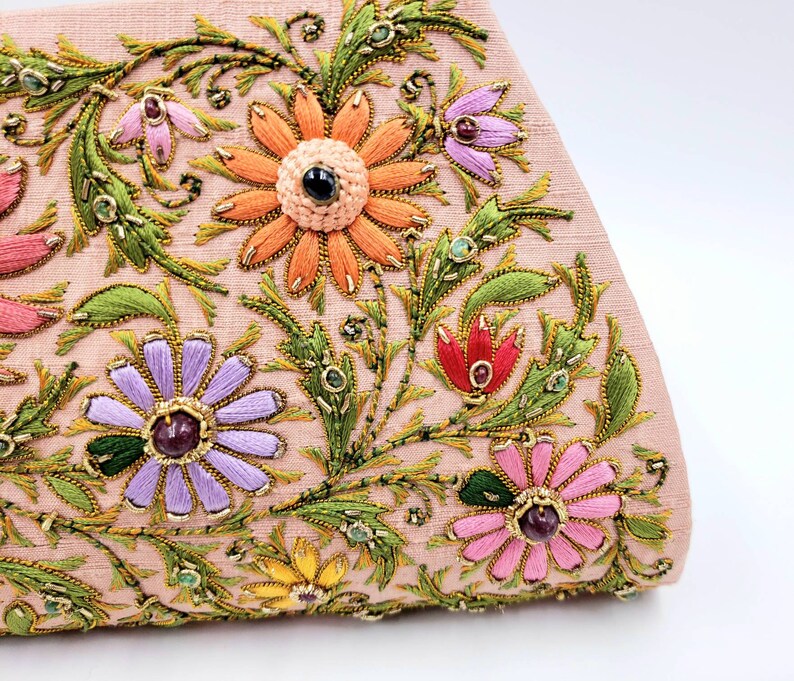 May include: A pink fabric clutch with embroidered floral designs in shades of green, orange, purple, pink, and red. The flowers are embellished with gold and jewel-toned beads.
