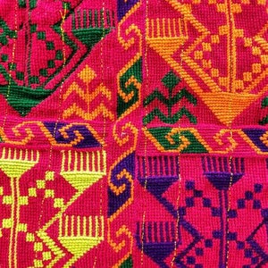 May include: Close-up of a vibrant textile with intricate embroidery. The design features geometric patterns in shades of red, green, yellow, orange, and purple. A black leather panel is at the top, with a silver and gold trim.