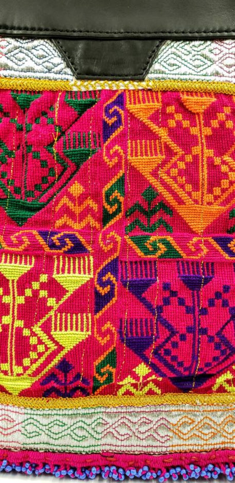 May include: Close-up of a vibrant textile with intricate embroidery. The design features geometric patterns in shades of red, green, yellow, orange, and purple. A black leather panel is at the top, with a silver and gold trim.