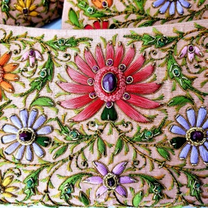 May include: A close-up of a pink fabric with intricate embroidery. The design features a large pink flower with a purple gem in the center, surrounded by smaller flowers and leaves. The fabric is decorated with gold thread and green and blue beads.