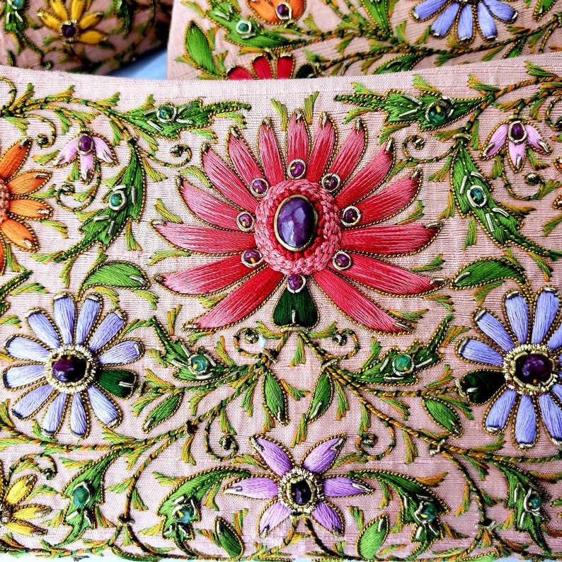 May include: A close-up of a pink fabric with intricate embroidery. The design features a large pink flower with a purple gem in the center, surrounded by smaller flowers and leaves. The fabric is decorated with gold thread and green and blue beads.