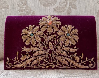 Könnte beinhalten: Eine bordeauxrote Samtclutch mit goldener Stickerei von drei Blumen und Blättern. Die Blumen haben grüne, orange und rosafarbene Edelsteine in der Mitte.