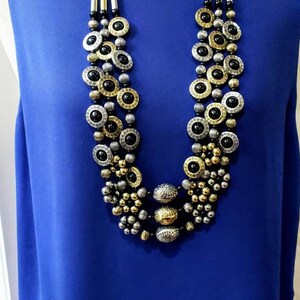 May include: A multi-strand necklace with black, gold, and silver round beads. The necklace is layered and features a large, ornate pendant.