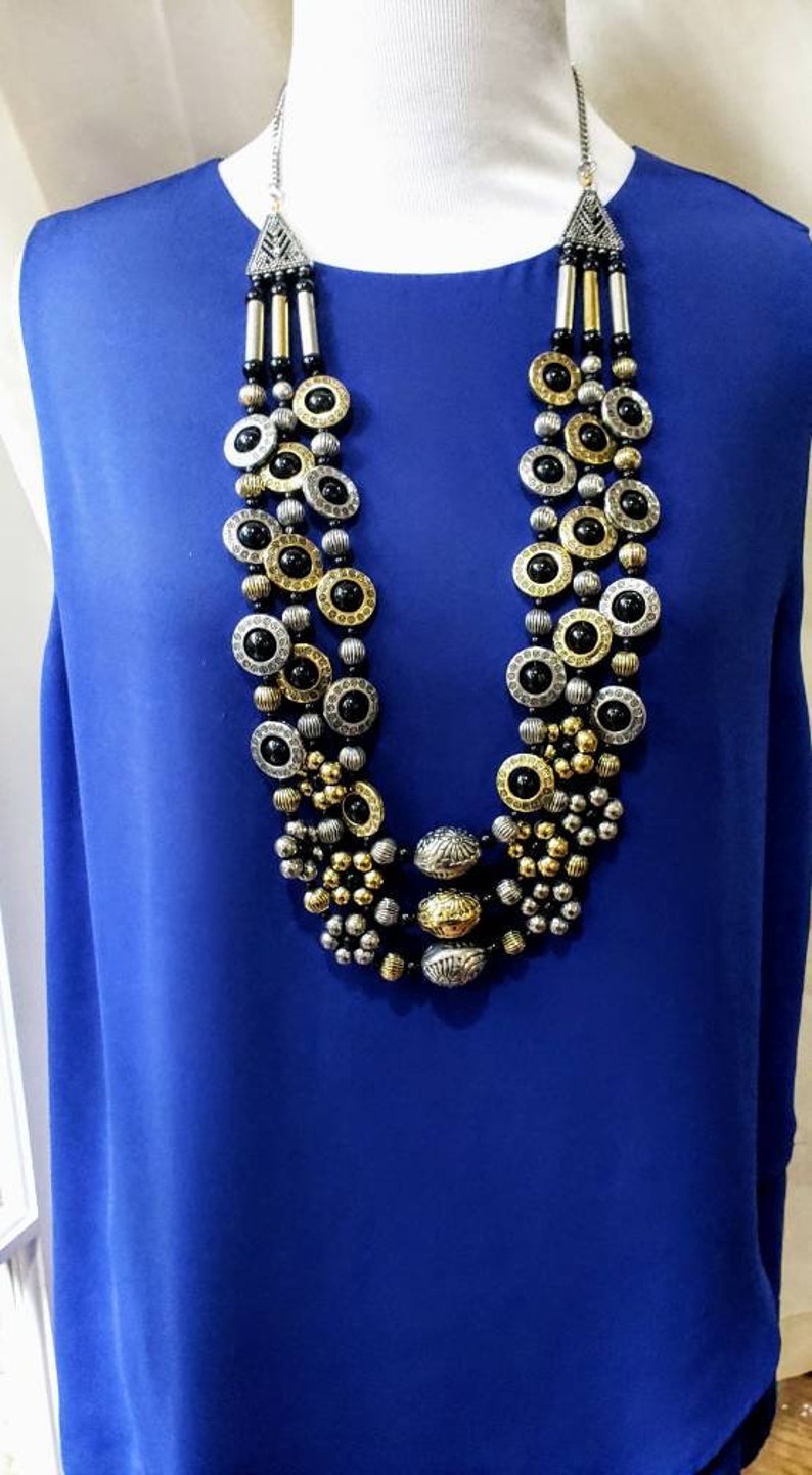 May include: A multi-strand necklace with black, gold, and silver round beads. The necklace is layered and features a large, ornate pendant.