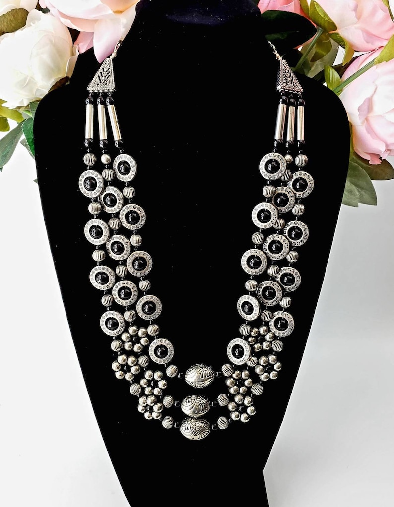 May include: A multi-strand necklace with silver-toned metal beads and black accents. The necklace features a variety of bead shapes, including round, oval, and cylindrical beads. The necklace is layered and has a decorative pendant at the center.
