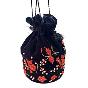 Op de afbeelding: Zwarte fluwelen trekkoordtas met rode bloemenborduursels en gouden kraalaccenten. De tas heeft een geplooid bovenstuk met een zwarte kwast en een trekkoordsluiting. Een stijlvol accessoire voor elke gelegenheid.