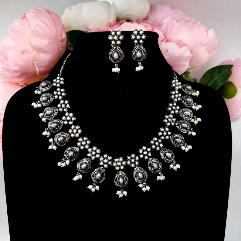 May include: A silver necklace and earrings set with a floral design and pearl accents. The necklace features a row of teardrop-shaped pendants with intricate details.