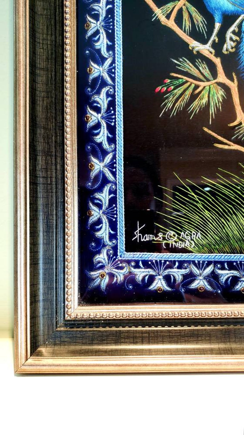 Puede incluir: Una obra de arte enmarcada que presenta un p&aacute;jaro azul posado en una rama con hojas verdes. El marco tiene un borde decorativo con dise&ntilde;os florales azules y blancos. La obra est&aacute; firmada "Khams Agra India".