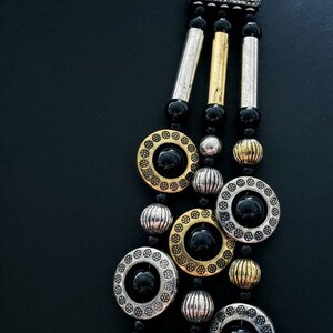 May include: A multi-strand necklace with black beads, gold and silver round charms, and a triangular pendant with black rhinestones.