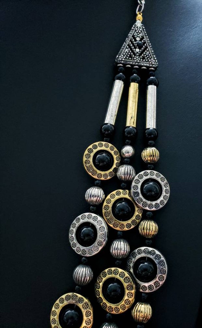 May include: A multi-strand necklace with black beads, gold and silver round charms, and a triangular pendant with black rhinestones.