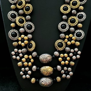 May include: A multi-strand necklace with black, gold, and silver beads. The necklace features large round beads with black centers and intricate designs. The necklace is layered with smaller beads in gold and silver.