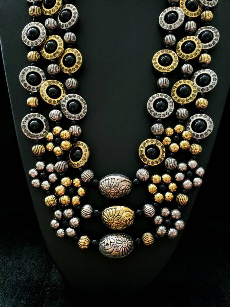 May include: A multi-strand necklace with black, gold, and silver beads. The necklace features large round beads with black centers and intricate designs. The necklace is layered with smaller beads in gold and silver.