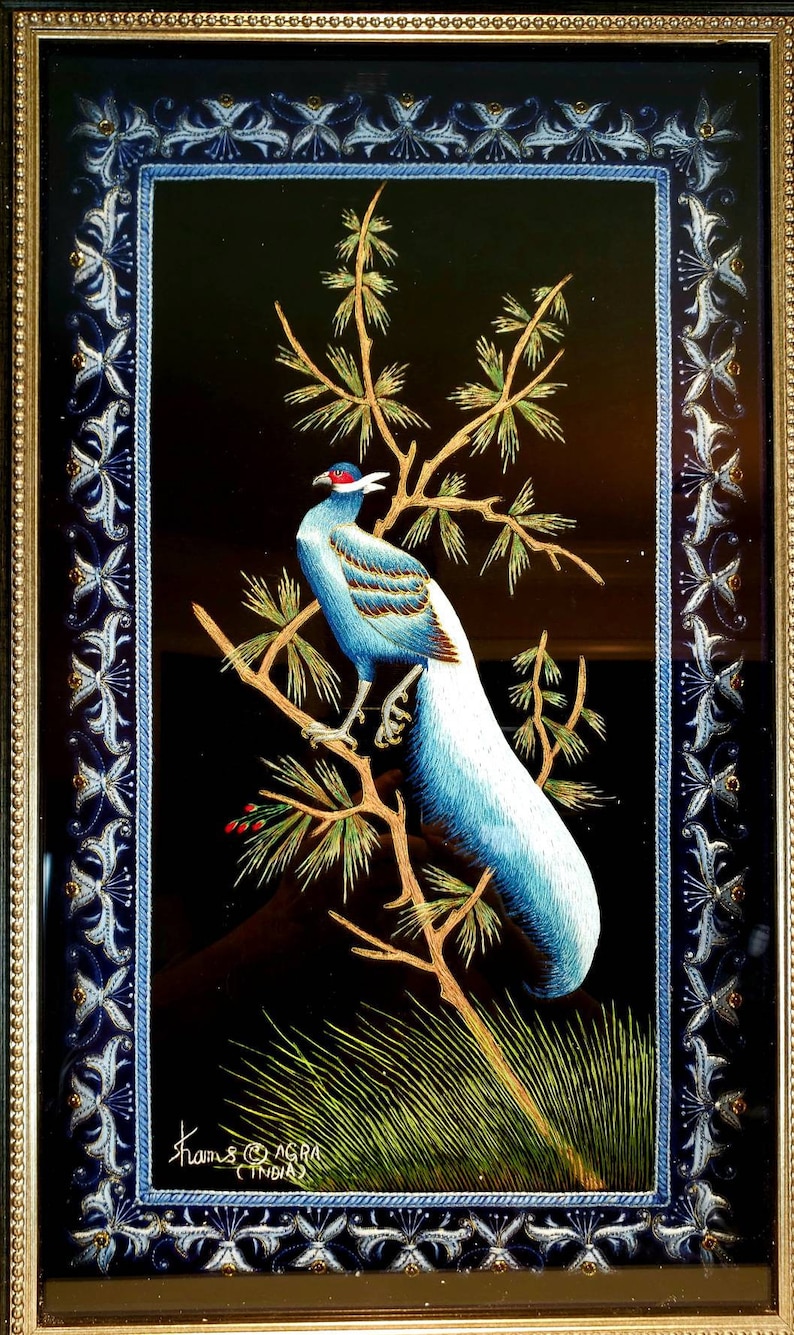 Puede incluir: Un bordado enmarcado que representa un fais&aacute;n azul y blanco posado en una rama de pino verde. El fondo es negro y el marco es dorado con un dise&ntilde;o floral azul y blanco. El bordado est&aacute; firmado "Shams Agra India".