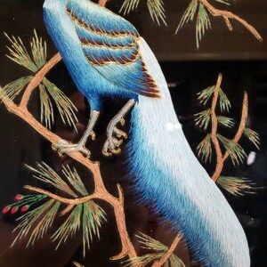 Puede incluir: Obra de arte bordada de un fais&aacute;n azul y blanco posado en una rama de pino con detalles dorados. El fondo es negro.