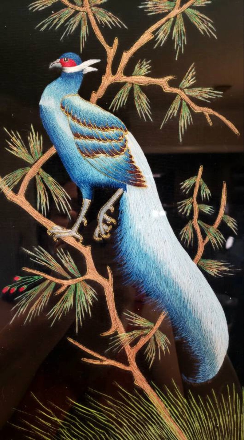 Puede incluir: Obra de arte bordada de un fais&aacute;n azul y blanco posado en una rama de pino con detalles dorados. El fondo es negro.