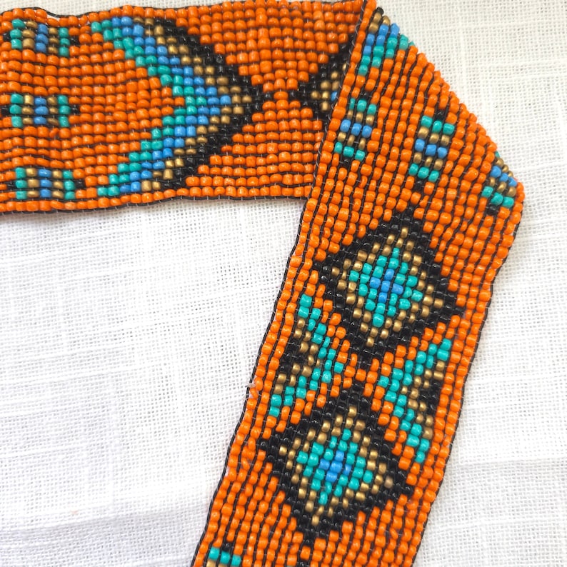 May include: A close-up of a beaded trim with a geometric pattern in orange, blue, black, and gold. The trim is made of small, round beads that are sewn together to create a repeating design.