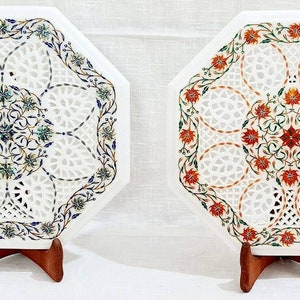 Puede incluir: Dos tapas de mesa octogonales de m&aacute;rmol blanco con intrincados dise&ntilde;os florales. Una tapa de mesa presenta acentos florales azules y verdes, mientras que la otra presenta acentos florales rojos y verdes. Ambas tapas de mesa est&aacute;n sobre soportes de madera.