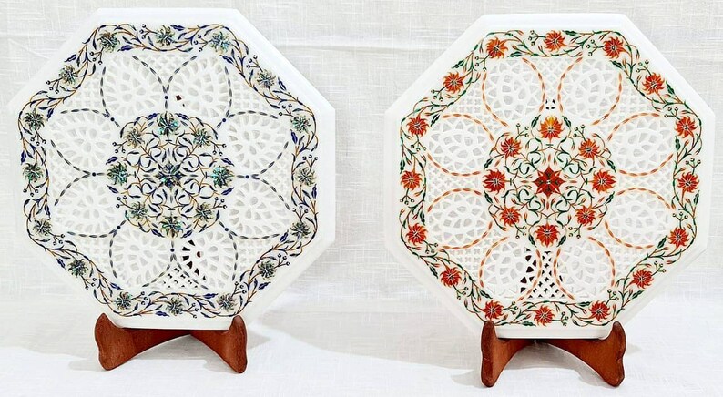 Puede incluir: Dos tapas de mesa octogonales de m&aacute;rmol blanco con intrincados dise&ntilde;os florales. Una tapa de mesa presenta acentos florales azules y verdes, mientras que la otra presenta acentos florales rojos y verdes. Ambas tapas de mesa est&aacute;n sobre soportes de madera.