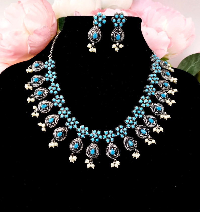 May include: A silver necklace and earrings set with turquoise blue stones and white pearl accents. The necklace features a teardrop design with multiple layers of turquoise stones.