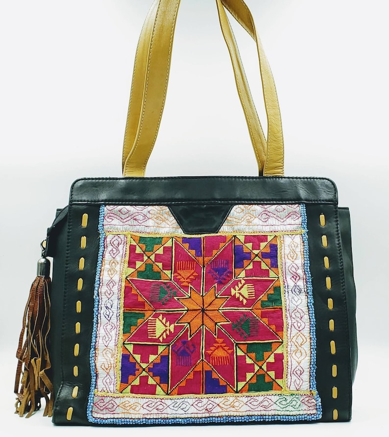 May include: Black leather tote bag with a colourful embroidered front panel featuring a starburst design in shades of pink, orange, green, and purple. The bag has tan leather handles and a brown tassel. The sides have decorative stitching.