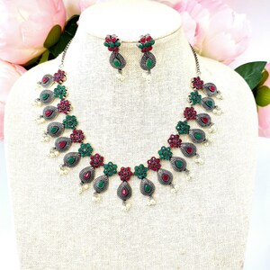 May include: A silver necklace and earrings set with red and green gemstone accents. The necklace features a teardrop design with pearl accents.